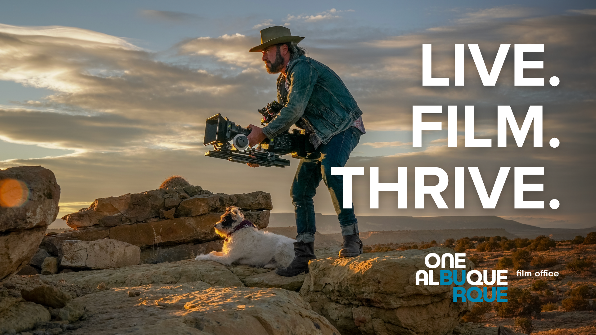 A cameraman in jeans and a denim jacket holds a camera mid body, looking into the distance in the colorful New Mexico desert, just outside of Albuquerque, at sunset with his terrier dog at his feet. The text "LIVE. FILM. THRIVE." is overlaid, with the Albuquerque Film Office logo.