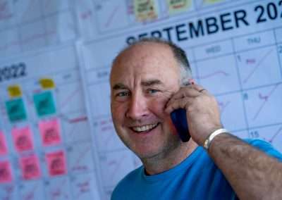 A man wearing a blue t-shirt smiles inot the camera while talking on a cell phone. The background is large calendars marked with information for work.