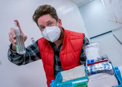 A man wearing a mask, a plaid shirt, and a red puffy vest looks into the camera while offering a bottle of hand sanitizer. He is in a hallway with white walls and floors, and is holding a tray with sanitizing supplies.