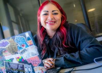 A woman with bright red and black hair smiles at the camera while sitting at a desk. In the foreground is her laptop, covered in stickers.