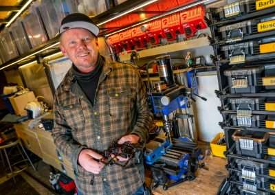 A man wearing a baseball hat and a plaid shirt smiles at the camera while holding a special effects tool with wires. In the background are industrial shelves and various tools.