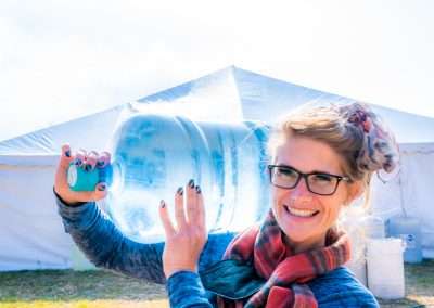 A woman with blonde hair tied up in a scarf, and glasses, smiles into the camera while she carries a large water jug over her shoulder. There is a large white tent in the background.