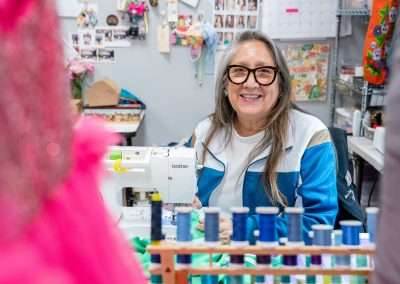 A woman with long gray hair and glasses smiles at the camera while sitting at a sewing machine. She is wearing a blue and white jacket, and the background is filled with colorful thread spools, fabric, and various decorations pinned to the wall. The foreground includes a blurred pink fabric, adding a pop of color to the scene.