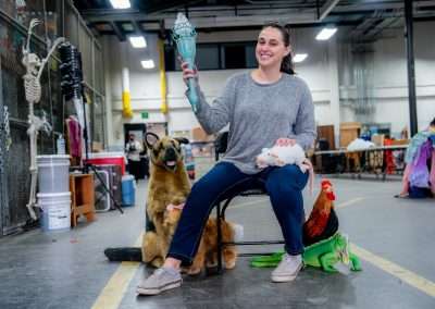 A woman wearing a gray sweater and jeans sits on a chair in a warehouse, holding a prop torch in one hand. She is surrounded by various animal props, including a German Shepherd, a white rat, a rooster, and an iguana. The background includes industrial shelves, containers, and other props.