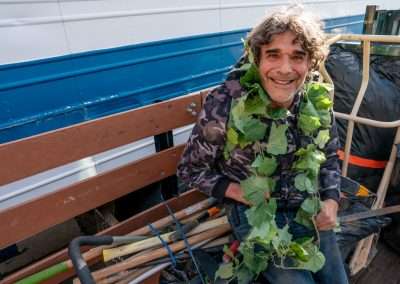 A man with curly hair, wearing a camouflage hoodie, smiles at the camera. He is sitting on a bench with green vines draped around his neck and shoulders. The background includes a blue and white wall and various gardening tools.