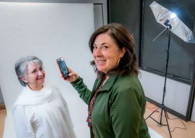 A woman looks into the camera, smiling, while taking a cell phone photo of another woman dressed all in white. They are in a photo studio with lighting equipment.