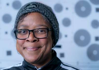 A photo of a woman looking into the camera with a closed-mouth smile. She is wearing a grey beanie and glasses.